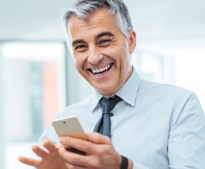 Smiling cheerful businessman using a touch screen smart phone and looking at camera