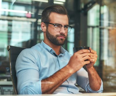Fresh coffee. Portrait of young and successful bearded man holding cup of coffee and looking at laptop while sitting at his working place. Business concept. Work concept